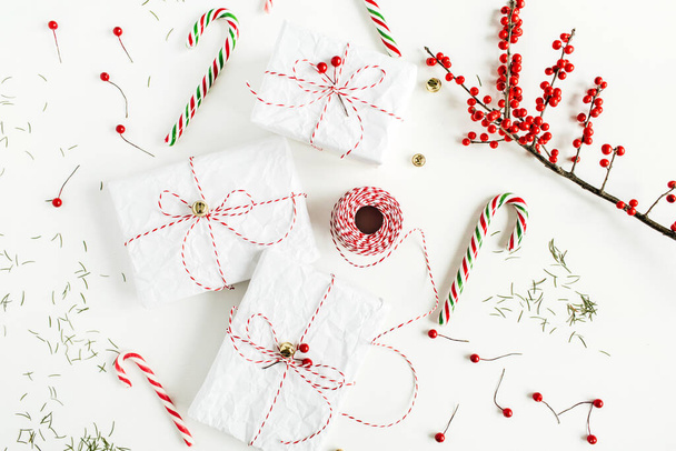 Karácsonyi ajándékdobozok ünnepi cukorkákkal, piros bogyós ágak, karácsonyi kötél tekercs és fenyő tűk fehér asztalon. Karácsony, újévi kompozíció. Lapos fekvésű, felülnézet.  - Fotó, kép