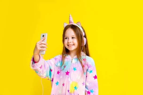Glückliches kleines Mädchen in einem Einhorn-Onesie, das Musik von seinem Handy hört und Spaß auf gelbem Hintergrund hat - Foto, Bild