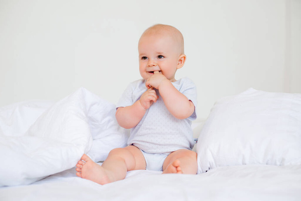 Φωτογραφία ενός χαρούμενου αγοριού 6 μηνών με λευκό κοστούμι στο κρεβάτι  - Φωτογραφία, εικόνα