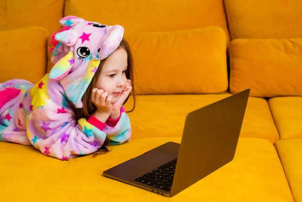 Klein meisje in eenhoorn onesie ligt thuis op een bank met een laptop. Geïsoleerd op oranje achtergrond, afstandsonderwijs voor kinderen en zelfisolatie - Foto, afbeelding