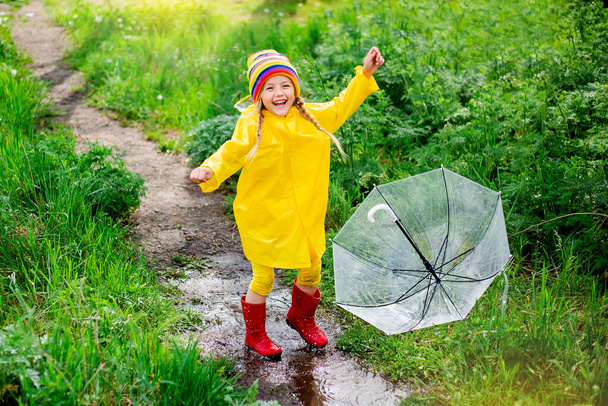 χαρούμενο κορίτσι με κίτρινο αδιάβροχο και διάφανη ομπρέλα πηδάει σε λασπωμένη λακκούβα - Φωτογραφία, εικόνα