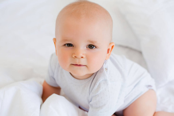6ヶ月の明るい赤ん坊の男の子の写真でベッドの中の白いボディスーツ  - 写真・画像