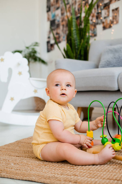 Cute toddler in yellow striped bodysuit sits playing with developmental toy on the carpet. Child development concept - Foto, imagen