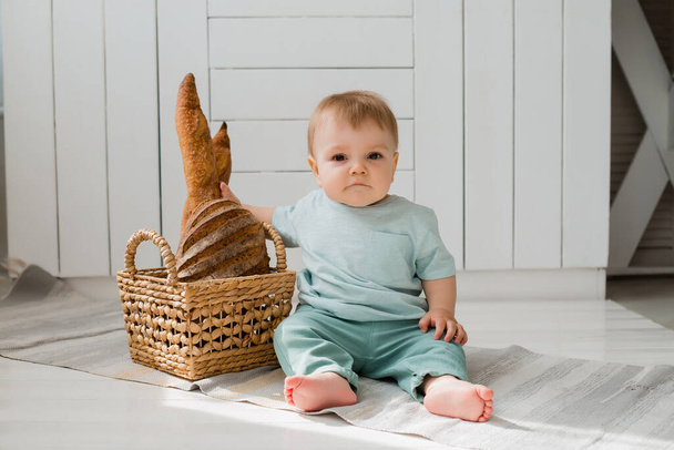 Mały chłopiec w miętowym kolorze ubrania siedzi w kuchni z koszem świeżego chleba - Zdjęcie, obraz