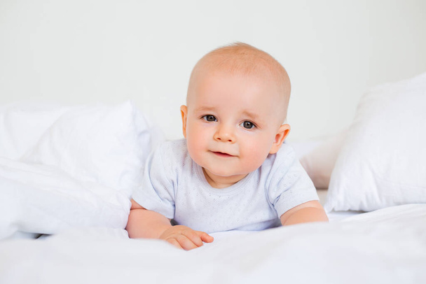 Photo of a cheerful baby boy of 6 months in a white bodysuit in bed  - Foto, imagen