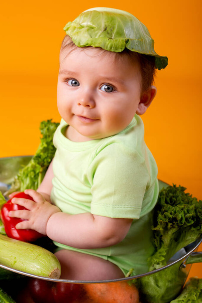 Хлопчик сидить у великій мисці зі свіжими овочами з капустяним листям, як його капелюх, концепцією екологічно чистих сільськогосподарських продуктів. Ізольоване зображення на помаранчевому фоні
 - Фото, зображення