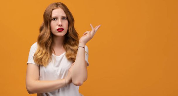 Studio shot of displeased girl with wavy redhead, wearing white t-shirt, gesturing with raised palm, frowning, being displeased and confused with dumb question. Human emotions concept. Copy space - Photo, image