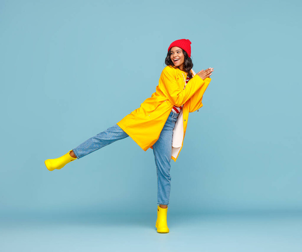 Full body cheerful ethnic woman in raincoat and boots smiling and kicking air on autumn day against blue backgroun - Photo, Image
