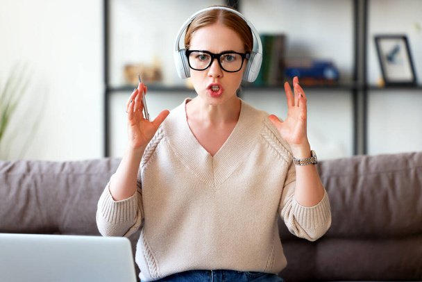 Smart adult female in headphones and glasses gesticulating and looking at camera while sitting on sofa and sharing wisdo - Фото, изображение