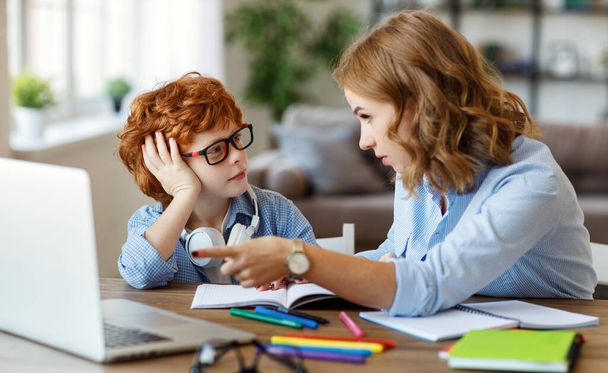Giovane madre che punta al computer portatile mentre spiega lezione al piccolo figlio in occhiali durante lo studio online insieme a hom - Foto, immagini