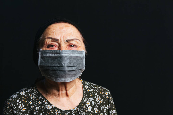 Retrato de uma mulher idosa em máscara médica em um fundo preto. Idosos em zona de risco por epidemia de vírus, pandemia de COVID-2019 - Foto, Imagem