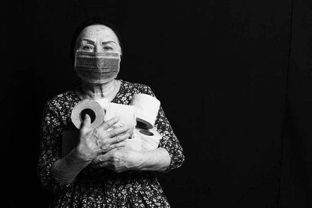 Czarno-biały portret starszej kobiety w masce medycznej trzymającej rolki papieru toaletowego na czarnym tle. COVID-2019 gorączka gromadząca - Zdjęcie, obraz