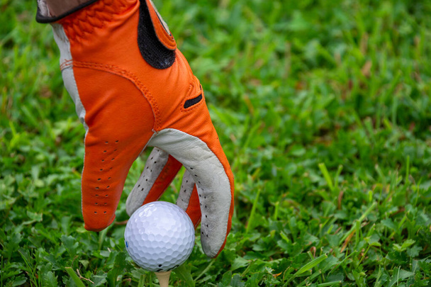 View of hand with orange glove and white golf ball on tee, on green grass, horizontal, with copy space - Photo, Image