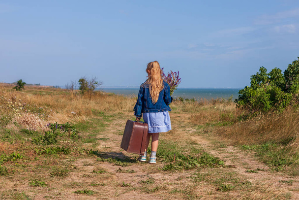 Little blonde girl in denim jacket, blue dress with vintage suitcase and flowers bouquet off-road with sea landscape. Stylish hitchhiker child with long hair on countryside trip. Kid walking outdoors. - Photo, Image