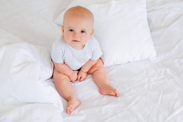 Φωτογραφία ενός χαρούμενου αγοριού 6 μηνών με λευκό κοστούμι στο κρεβάτι  - Φωτογραφία, εικόνα