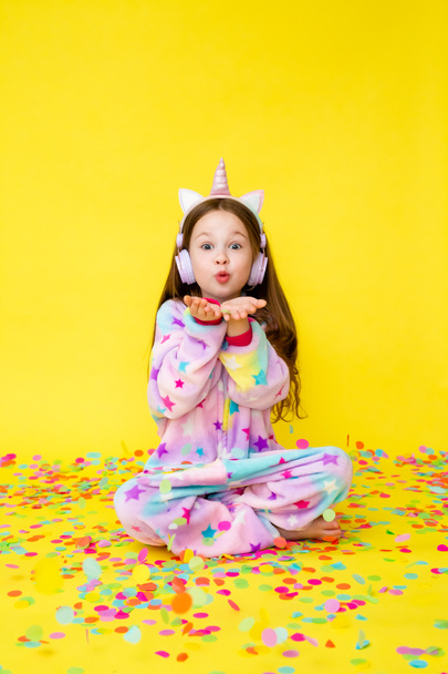 Klein meisje met lang haar in eenhoorn jumpsuit en oortjes spelen met kleurrijke confetti op een gele achtergrond. Kopieer ruimte foto voor uw advertentie - Foto, afbeelding