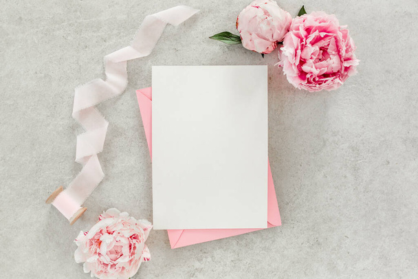 Mockup invitation, blank paper greeting card, pink envelope and peonies on gray stone table. Flower background. Flat lay, top view. - Фото, изображение