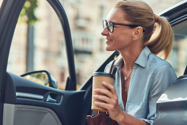 Onnellinen menestyvä keski-ikäinen liikenainen, joka pitelee kahvikuppia noustessaan autostaan, saapuu töihin - Valokuva, kuva