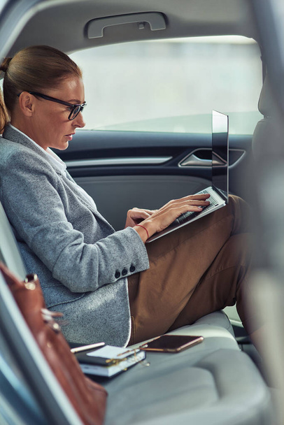 Working online. Side view of a busy middle aged businesswoman using laptop while sitting on back seat in the car - Fotó, kép