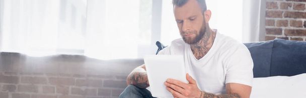 vízszintes kép szakállas tetovált férfi segítségével digitális tabletta a hálószobában - Fotó, kép