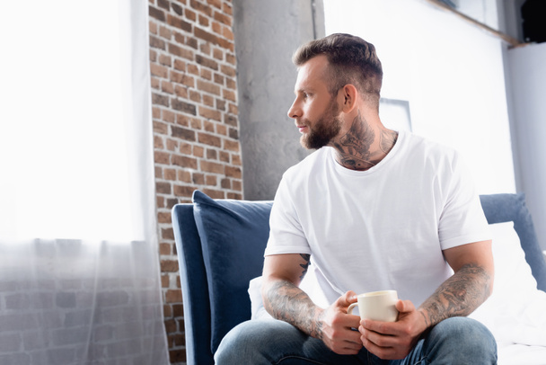 przemyślany mężczyzna w białej koszulce spoglądający przez okno siedząc na łóżku z filiżanką herbaty - Zdjęcie, obraz