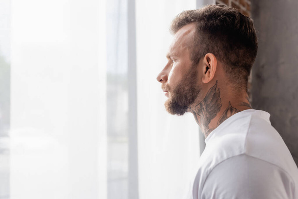 vista lateral del hombre barbudo y tatuado mirando hacia otro lado mientras está de pie cerca de la ventana en casa - Foto, imagen