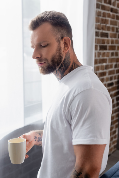 thoughtful bearded man in white t-shirt holding cup of tea while standing near window - Φωτογραφία, εικόνα