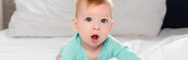 horizontal image of infant boy looking at camera while crawling in bed with open mouth - Photo, Image