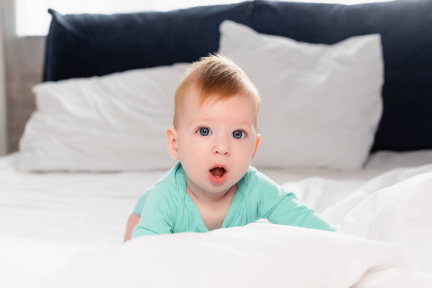 enfoque selectivo del niño mirando a la cámara con la boca abierta mientras se arrastra en la cama - Foto, Imagen