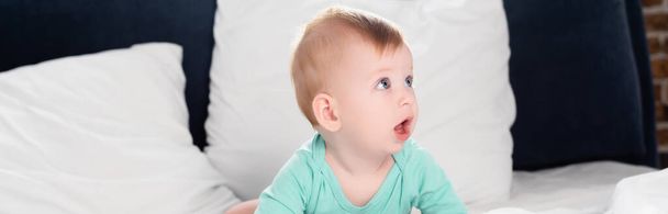 panoramic crop of infant boy looking away while crawling in bed with open mouth - Photo, Image