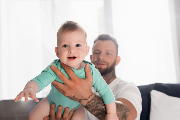 selective focus of excited baby boy in hands of young father in bedroom - Photo, Image
