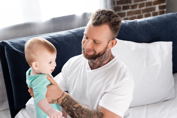 young father in white t-shirt holding infant child while sitting on bed - Photo, Image