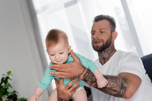 szelektív fókusz szakállas tetovált férfi holding baby boy a hálószobában - Fotó, kép