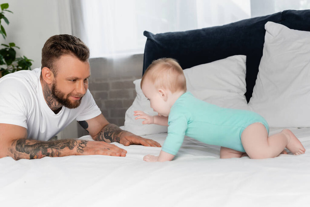 selective focus of tattooed young man having fun with infant boy crawling on bed in baby romper - Photo, Image