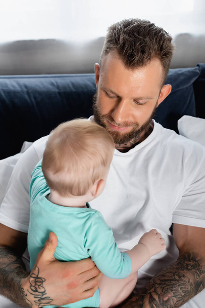 young tattooed father holding baby boy while sitting in bedroom - Foto, Imagem