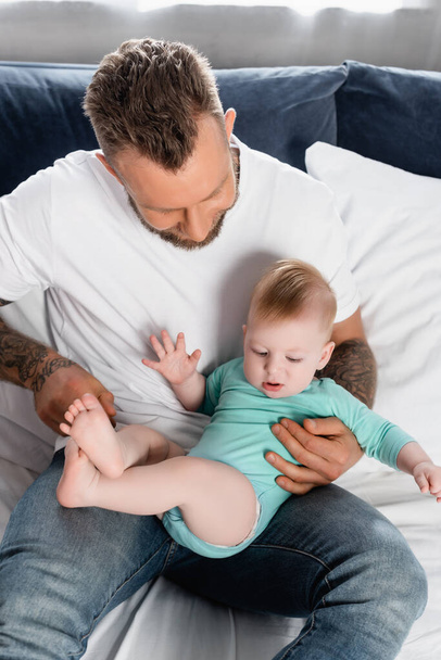 young father in white t-shirt and jeans sitting on bed and having fun with infant son - Photo, Image