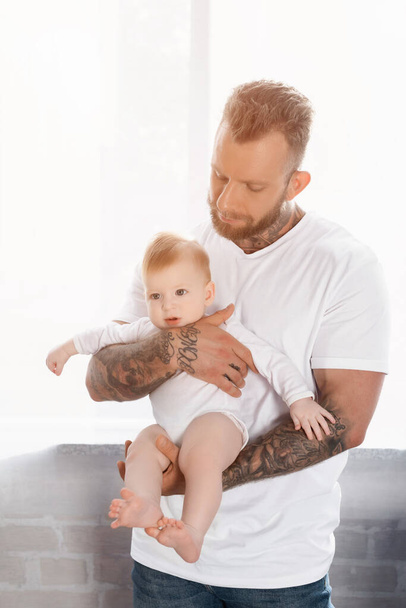 młody brodaty mężczyzna w białym t-shirt gospodarstwa niemowlę dziecko w baby romper - Zdjęcie, obraz
