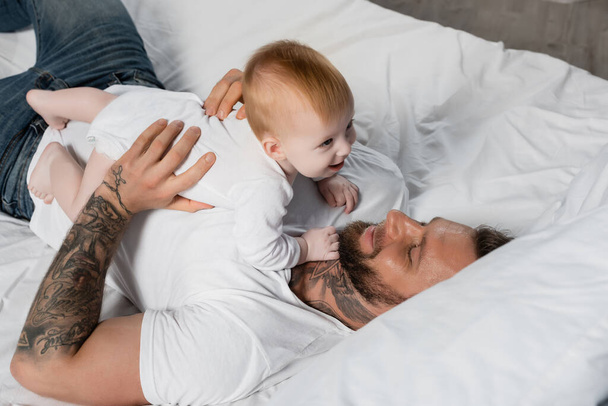 overhead view of young tattooed man having fun with baby boy while lying in bed - Photo, Image