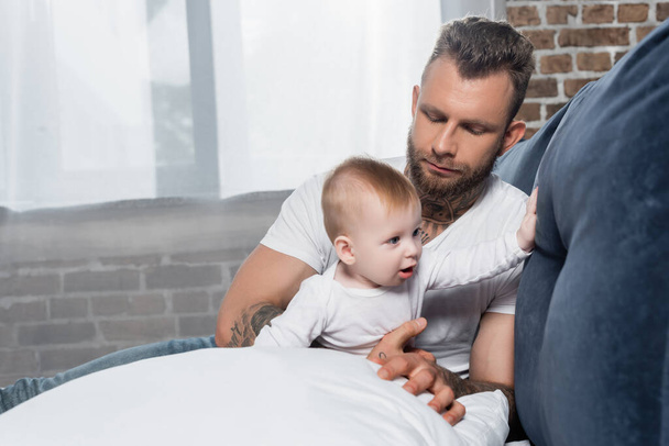 selective focus of young bearded man sitting on bed near infant child - Foto, afbeelding