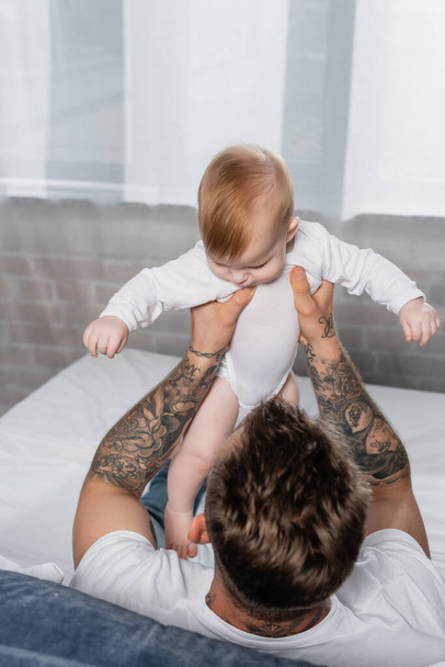 γενικά άποψη του άντρα με τατουάζ που κρατά το παιδί βρέφος στο μωρό romper ενώ κάθεται στο κρεβάτι - Φωτογραφία, εικόνα