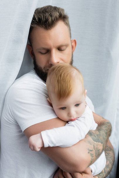 fiatal tetovált férfi fehér pólóban kezében csecsemő fia kezében csukott szemmel - Fotó, kép