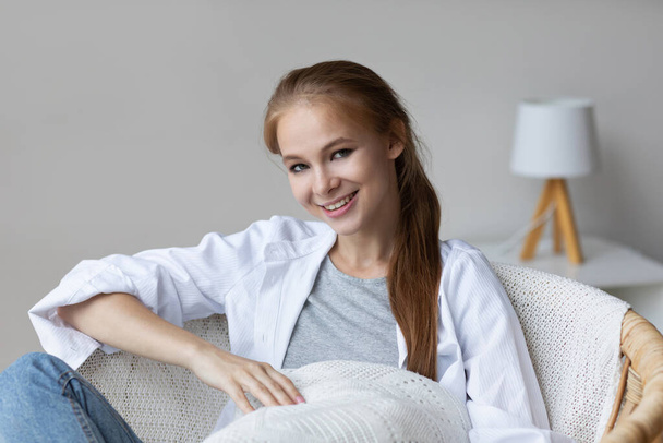 Beautiful happy woman looking at camera and smiling while sitting in a cozy chair. Female portrait indoors. - Zdjęcie, obraz