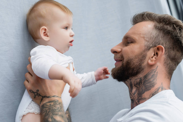 side view of joyful tattooed man holding baby boy in hands - Zdjęcie, obraz