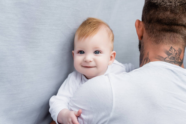 back view of tattooed man holding joyful infant child looking at camera - Photo, Image