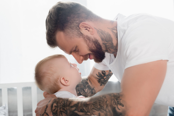 side view of joyful tattooed man and baby boy looking at each other face to face - 写真・画像