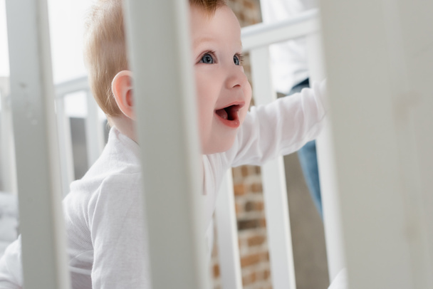selective focus of excited infant boy with open mouth in crib - Foto, afbeelding