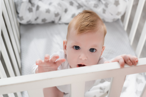 selective focus of infant boy looking at camera while standing in crib with outstretched hand - Foto, Imagen