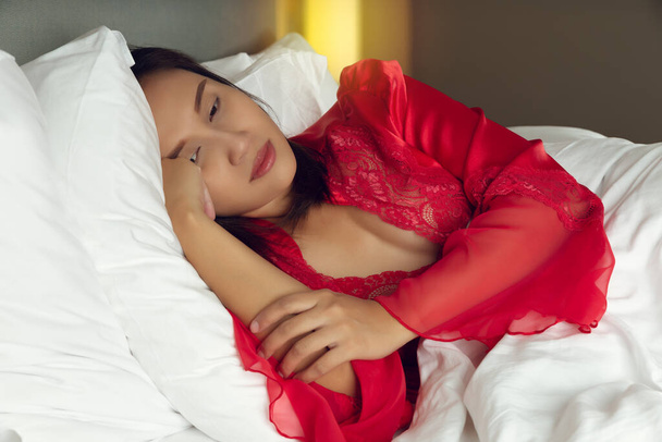 Vermoeide vrouw in bed kan 's nachts niet slapen met slapeloosheid. Aziatisch meisje droeg een satijnen nachtjapon en rode badjas liggend op het bed wakker in de slaapkamer - Foto, afbeelding