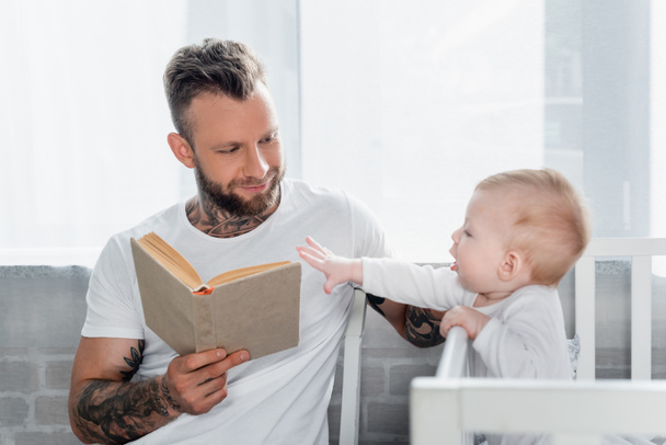 tattooed man reading book to baby boy standing in cot with outstretched hand - Photo, Image