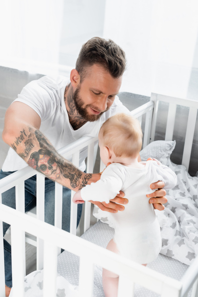 selective focus of young tattooed man supporting infant son standing in crib - Foto, Imagen